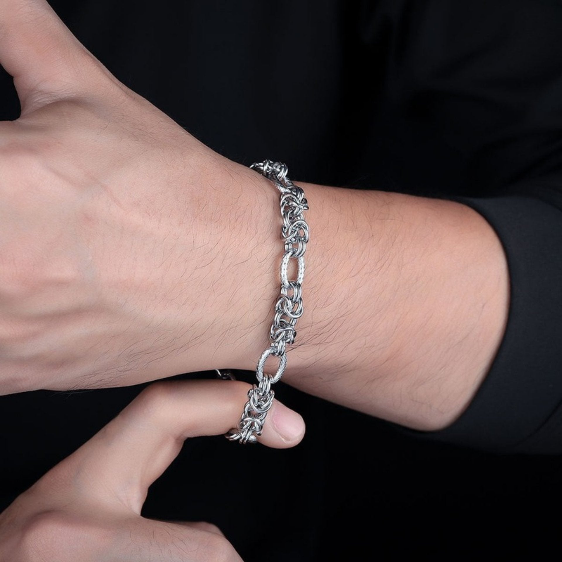 Close-up of a man’s wrist wearing a silver rope-style chain bracelet, with his thumb pointing up, against a black background.