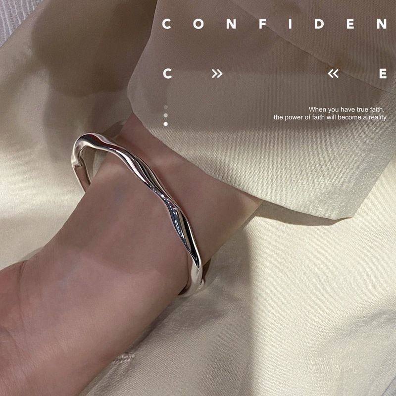 A model is wearing a silver wavy bangle bracelet on their wrist against cream silk fabric with inspirational text overlay that reads 'CONFIDENCE' and a quote about faith