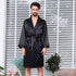 Man standing against white venetian blinds wearing a black silk robe with shawl collar and waist tie, surrounded by small potted plants and decorative elements