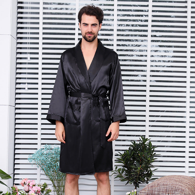 Man standing against white venetian blinds wearing a black silk robe with shawl collar and waist tie, surrounded by small potted plants and decorative elements