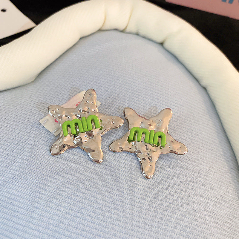 Silver star-shaped hair clips with a textured metallic finish and green "min" lettering, displayed on soft fabric.