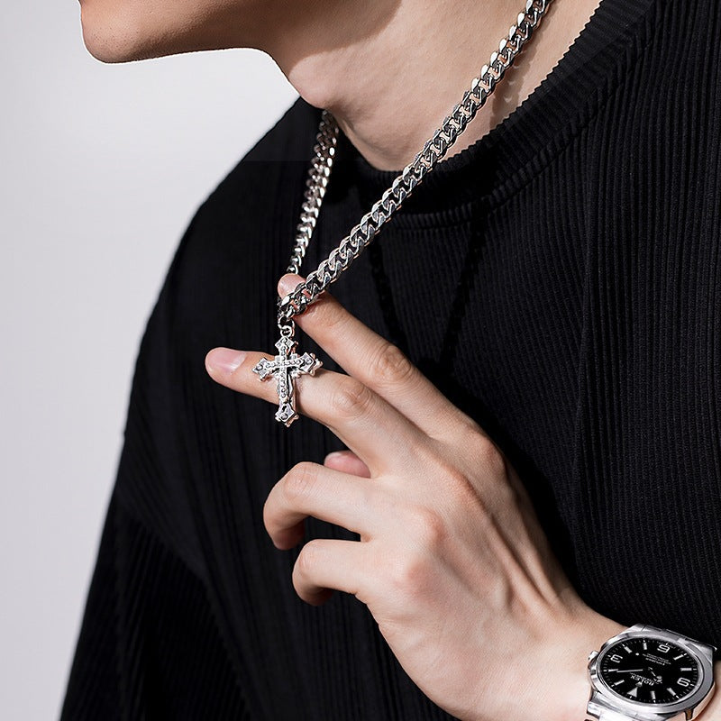 Man wearing a silver cross pendant necklace with a thick Cuban chain, holding the pendant against a black ribbed sweater