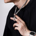 Man wearing a silver cross pendant necklace with a thick Cuban chain, holding the pendant against a black ribbed sweater
