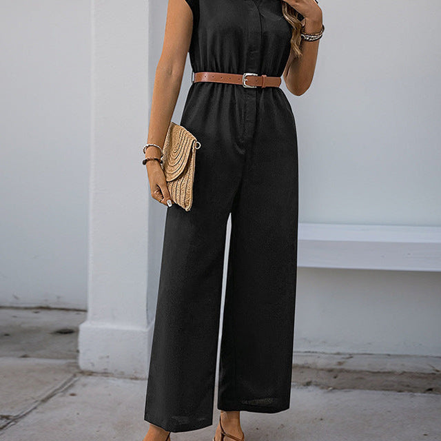 A woman in a black wide-leg jumpsuit with a tan belt, accessorized with a woven clutch and sandals, standing in an outdoor setting.