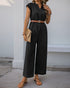 A woman in a black wide-leg jumpsuit with a tan belt, accessorized with a woven clutch and sandals, standing in an outdoor setting.
