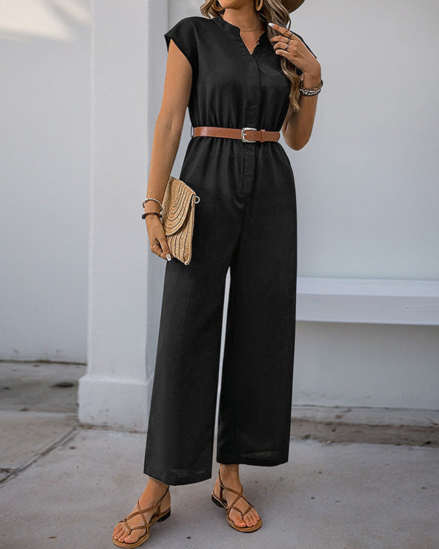 A woman in a black wide-leg jumpsuit with a tan belt, accessorized with a woven clutch and sandals, standing in an outdoor setting.