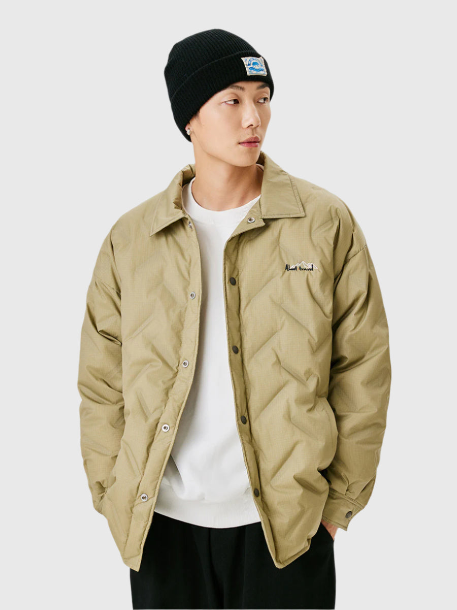 A man wearing a black ribbed beanie and tan quilted shirt jacket with embroidered logo, posing against white background