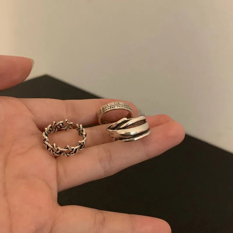 A person holding three silver rings in their palm - a Roman numeral ring, braided chain ring, and layered band ring against a neutral background