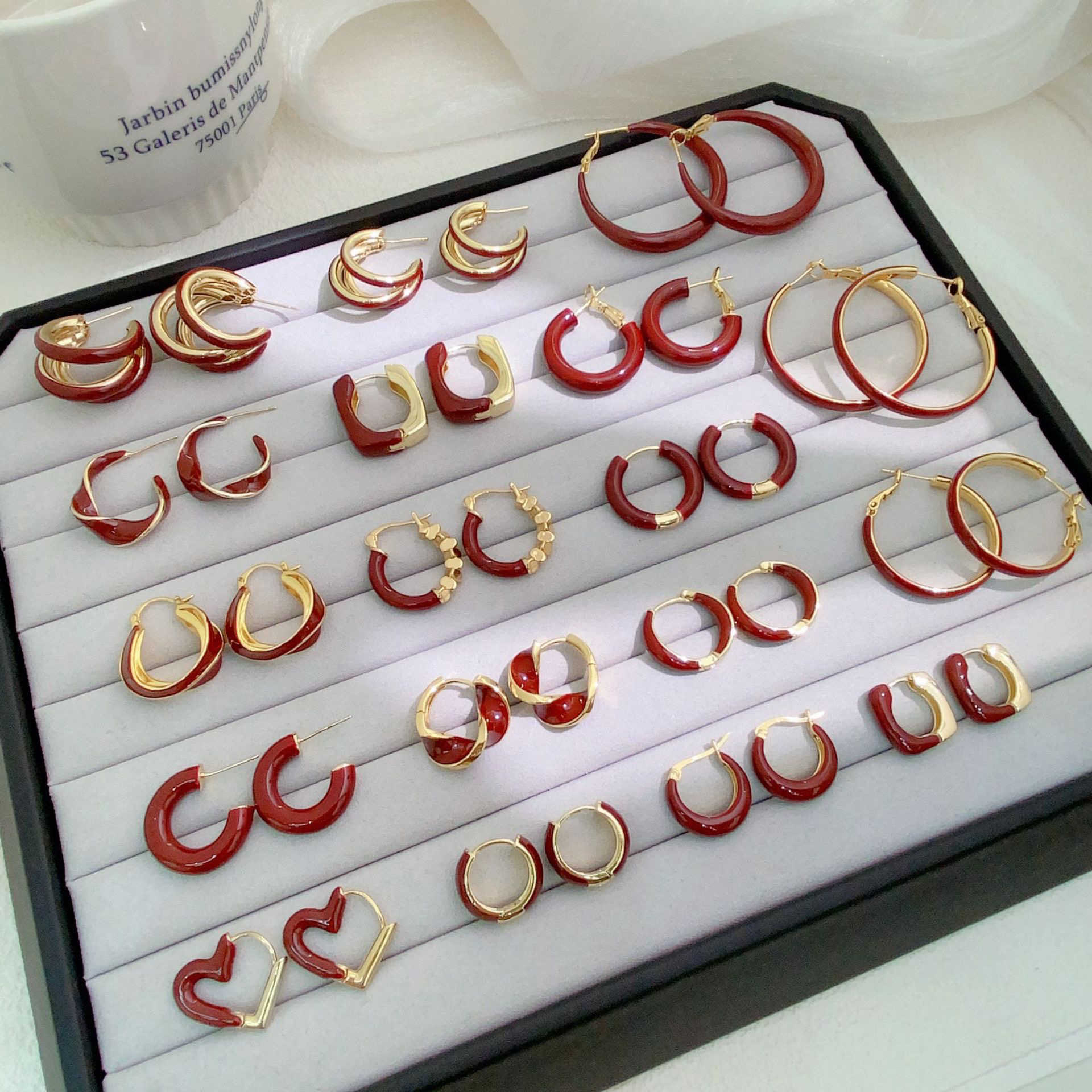 A jewelry display case features multiple pairs of gold and red enamel hoop earrings in various sizes and styles, arranged on a soft white cushion
