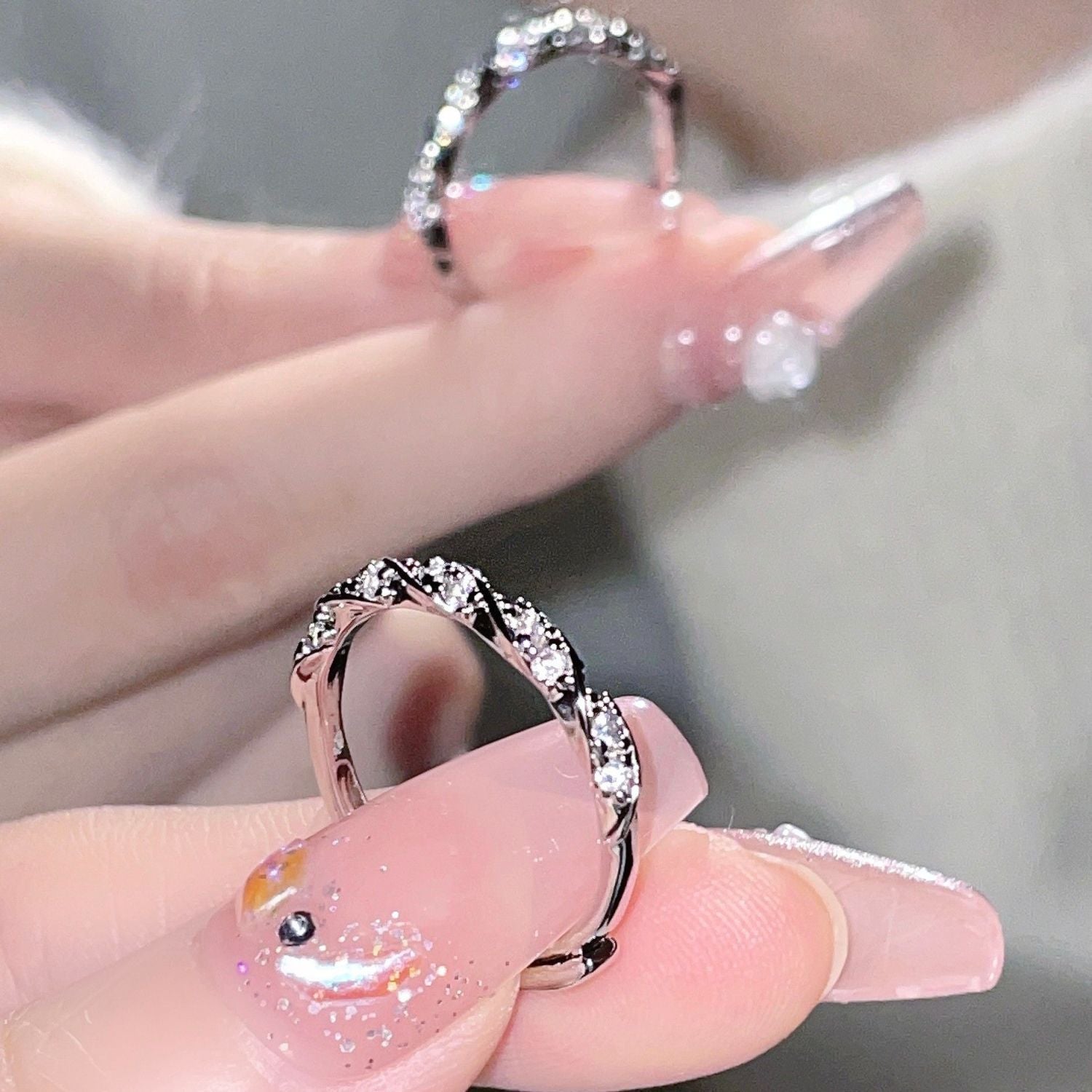 Close-up of hand with glitter pink nails displaying twisted diamond ring from two angles on light background