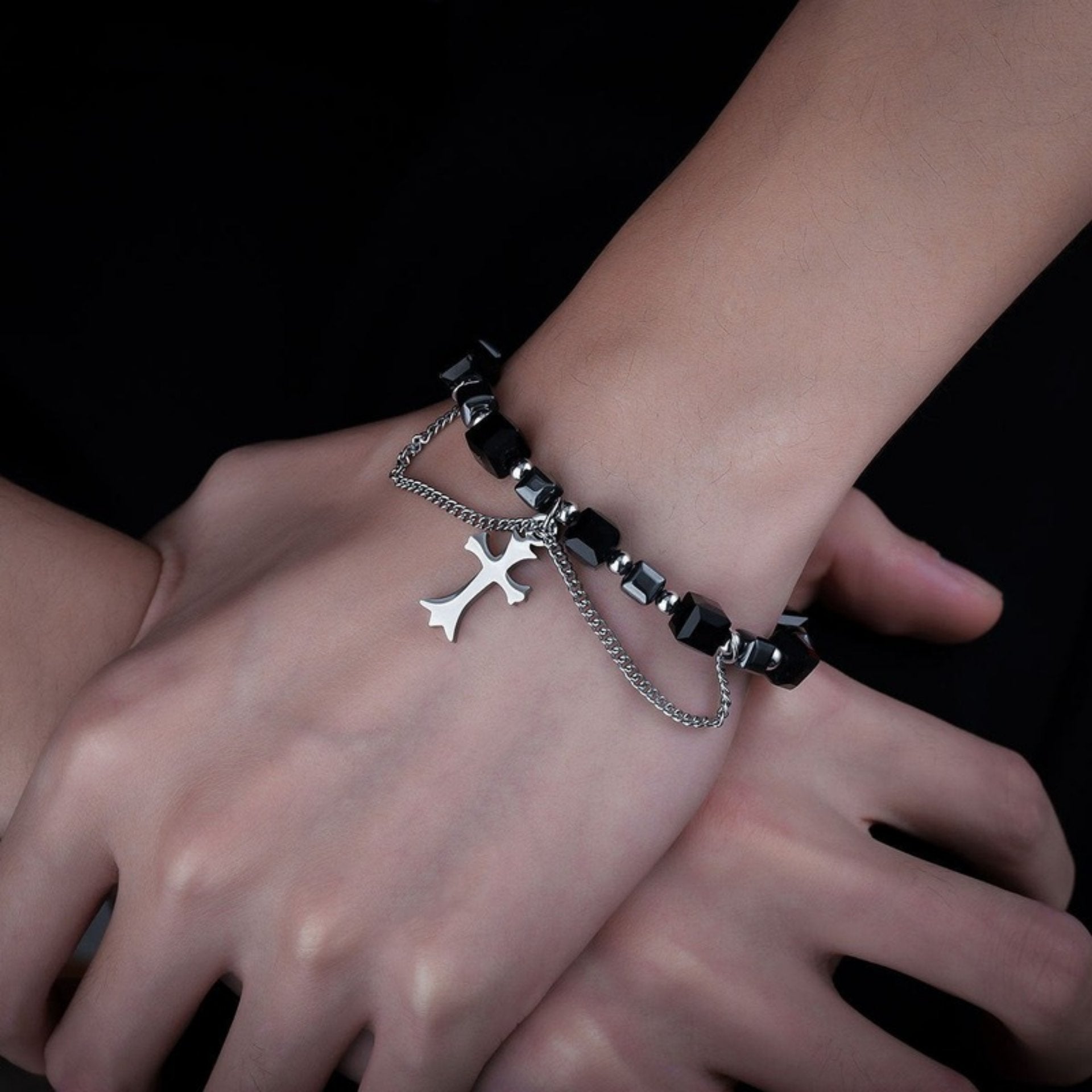 A close-up of a person's wrist wearing a black beaded bracelet with silver accents and a dangling silver cross pendant, styled with a chain detail.