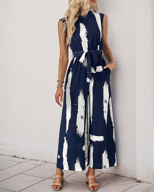 Woman wearing a navy sleeveless jumpsuit with white brushstroke print, a belted waist, and wide-leg pants, styled with neutral sandals.
