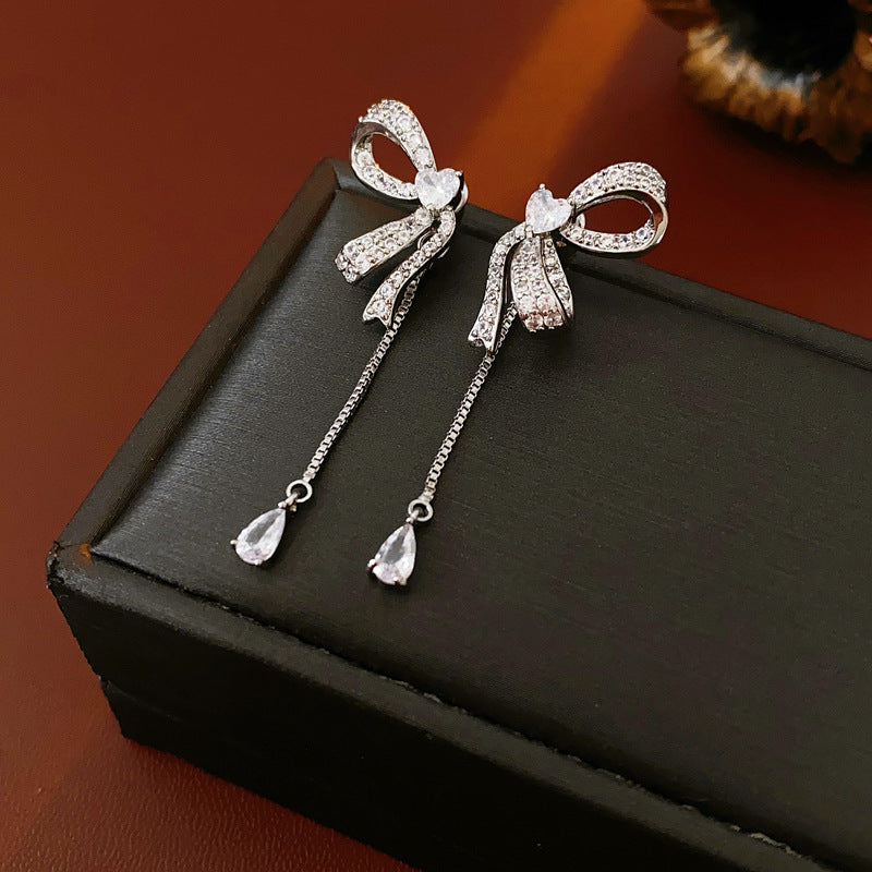 Crystal-adorned bow earrings with teardrop pendants displayed in white cushioned jewelry box