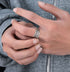 A close-up of a hand wearing a silver feather-design ring on the ring finger, against a black background.