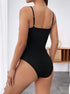 A woman wearing a black ribbed bodysuit with adjustable straps shown from behind, revealing the fitted silhouette against a white wall with wooden furniture