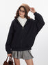 A model wears a black oversized zip-up hoodie, white shirt, gray pleated skirt, and a brown beanie, adjusting her hat while posing