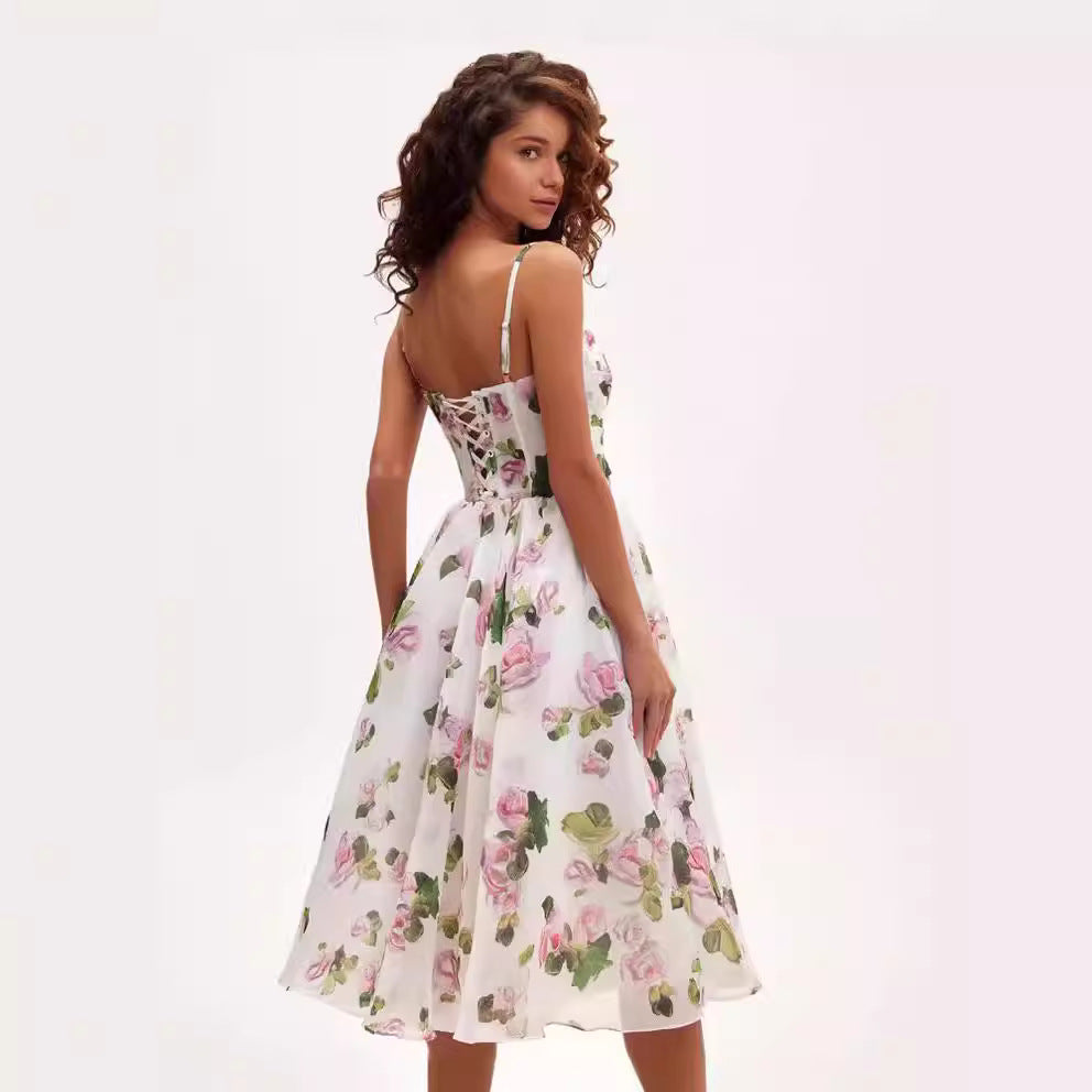 A model with curly hair is showing the corset-back detail of a white floral sundress with pink roses and green leaves pattern against white backdrop