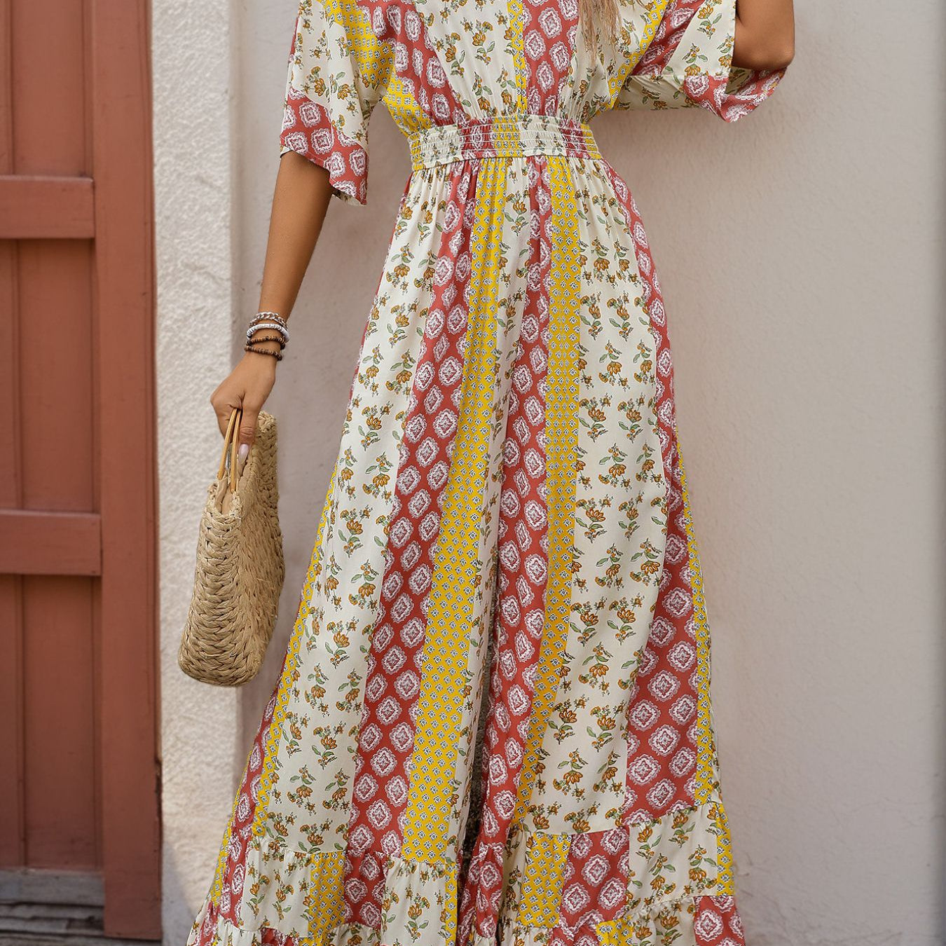 Woman in a bohemian jumpsuit with patchwork prints in red, yellow, and white, carrying a woven handbag and wearing sandals.