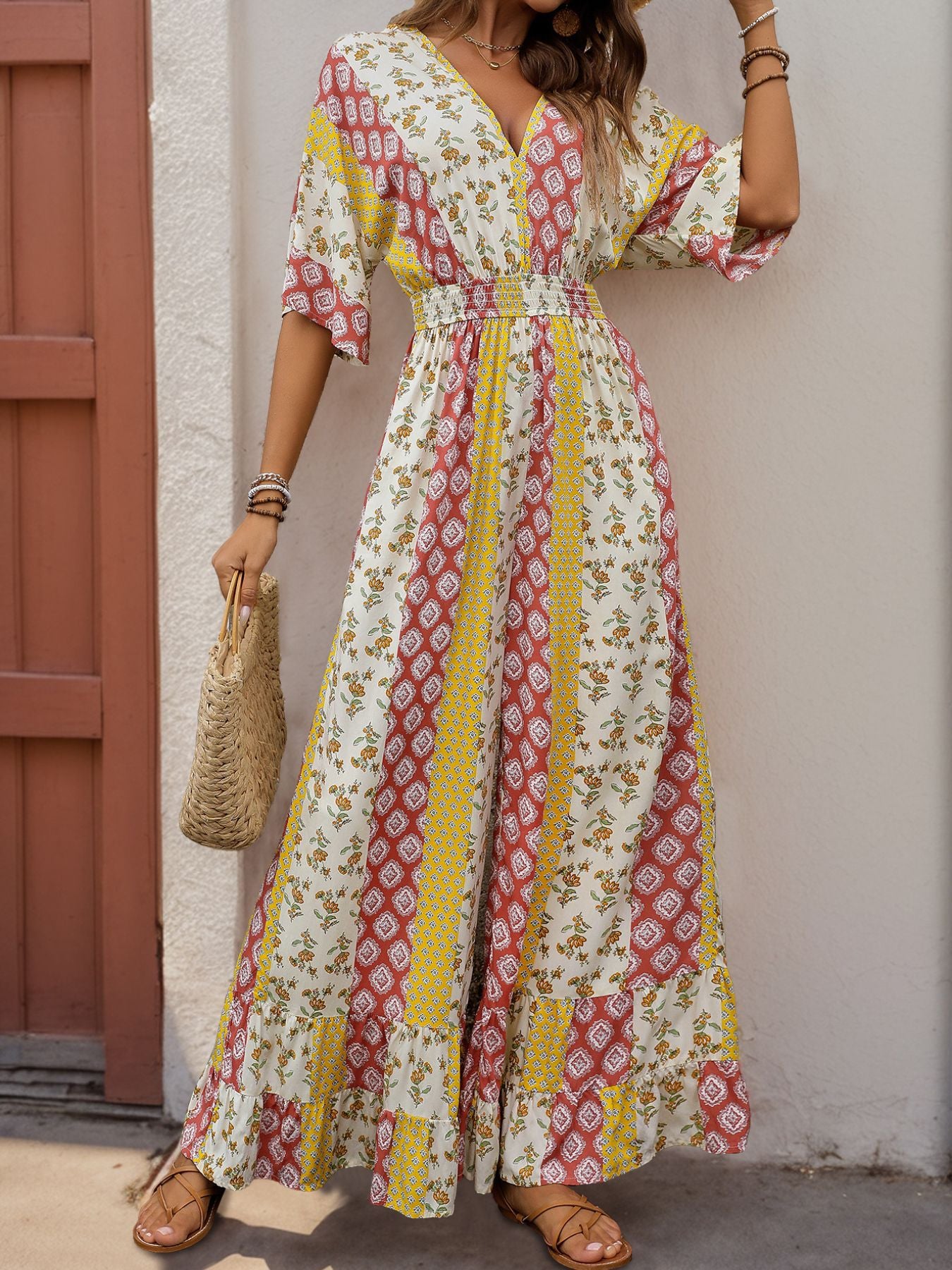 Woman in a bohemian jumpsuit with patchwork prints in red, yellow, and white, carrying a woven handbag and wearing sandals.