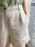 Close-up detail of beige linen shorts showing front pleats, side pocket, and brass button closure against light background