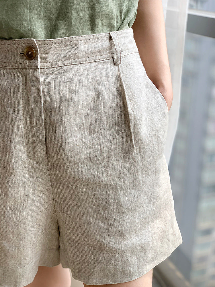 Close-up detail of beige linen shorts showing front pleats, side pocket, and brass button closure against light background