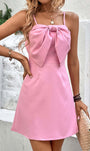 Pink bow front mini dress with spaghetti straps, fitted waist, and A-line skirt. Styled with layered necklaces, bracelets, and a woven clutch.