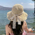 A woman wearing a beige straw sun hat with a ruffled brim and delicate lace ribbon, enjoying a scenic lakeside view.