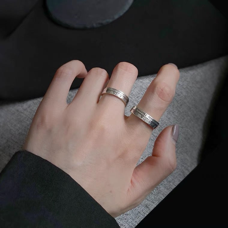 A person wearing silver adjustable Roman numeral rings on two fingers, showcasing vintage style jewelry against grey fabric