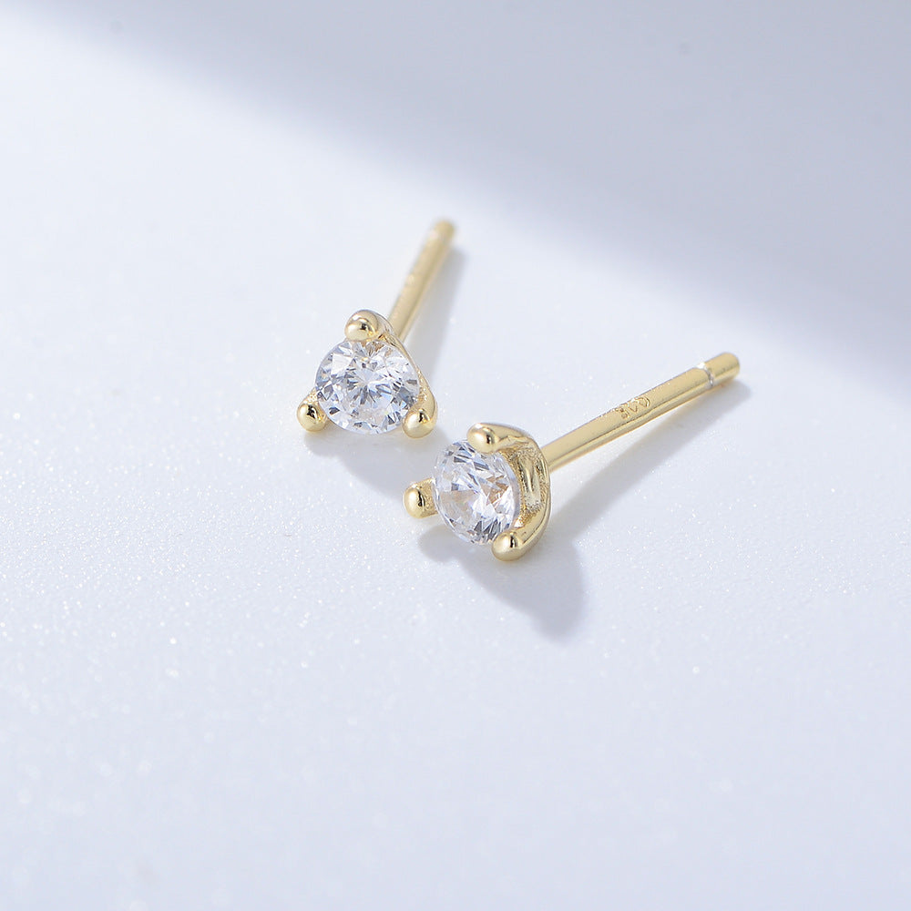 Gold three-prong diamond stud earrings with small round brilliant stones on white surface casting soft shadow