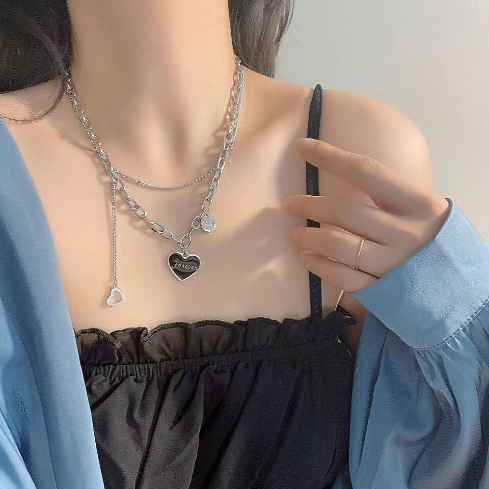 Layered silver chain necklace with "Forever" black heart pendant and small heart charm, worn with blue top