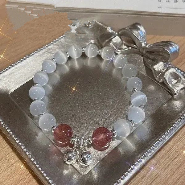 A gemstone bracelet with clear quartz beads and pink accent stones displayed on a silver decorative tray. Features small silver bell charms