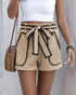Woman wearing beige high-waisted shorts with black lace trim and a matching belt, styled with a white crop top and accessories.