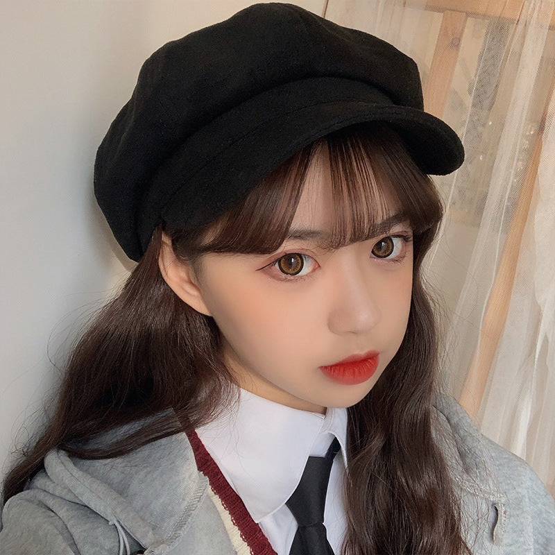 A woman wearing a black newsboy cap with white collar shirt and black tie, posing indoors against a light curtain background