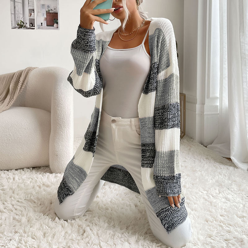 A woman sitting on white shag rug taking mirror selfie in open front striped cardigan, white bodysuit and white jeans with chain necklace detail