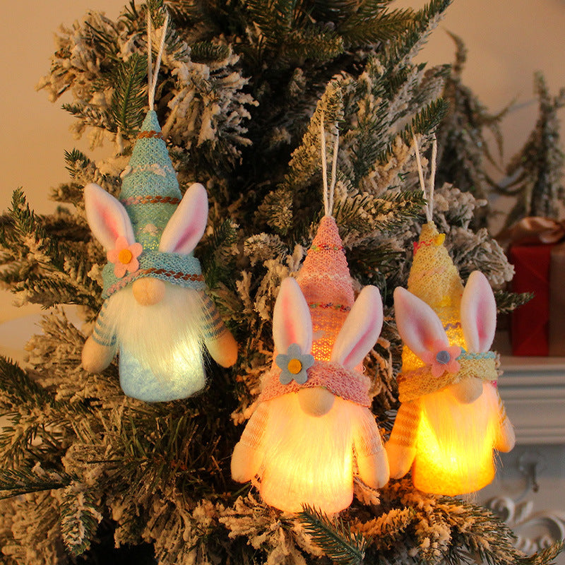 Three glowing bunny gnome ornaments with pastel hats and floral details, hanging on a snow-covered Christmas tree.