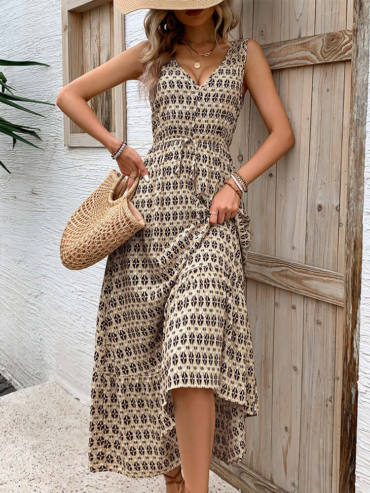  Woman wearing a beige and black printed V-neck sleeveless maxi dress with a cinched waist, accessorized with a woven bag and gold jewelry.
