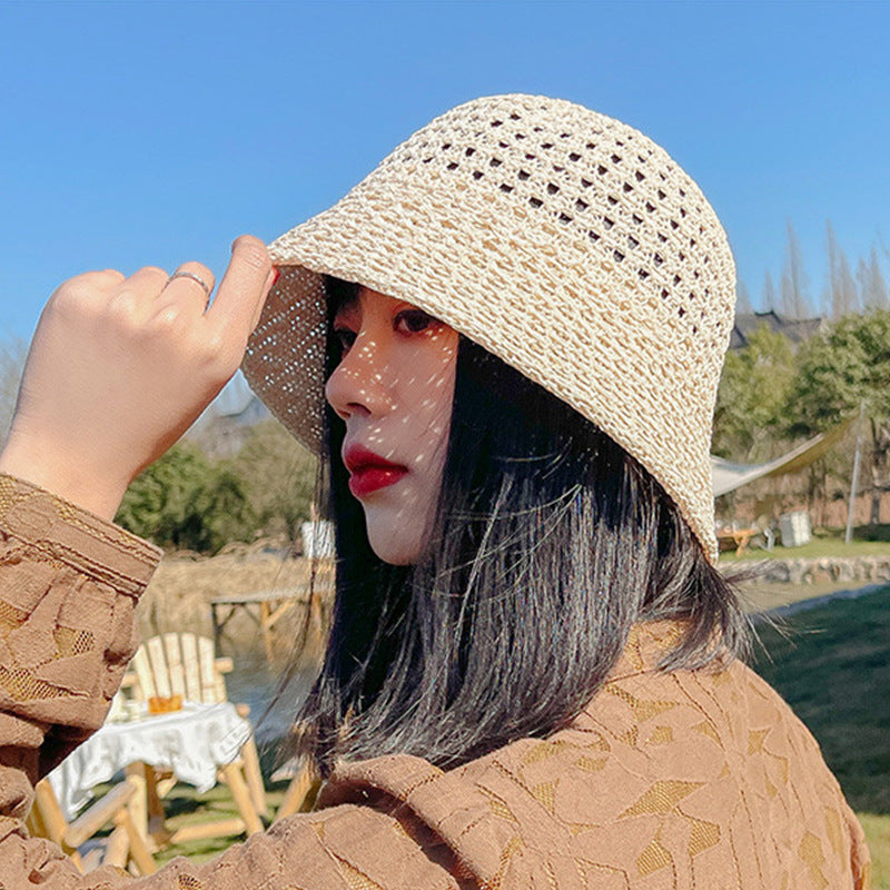 A woman wearing a beige woven bucket hat with an open-weave pattern, casting shadows on her face, paired with a brown textured blouse against a sunny outdoor backdrop.