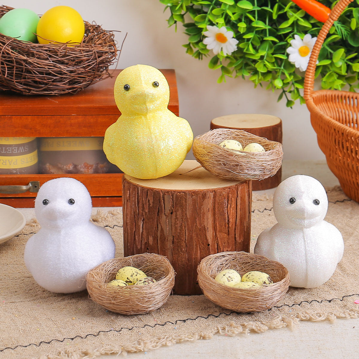Glittery yellow and white chick figurines with black eyes displayed on wooden logs, accompanied by small nests with speckled eggs.