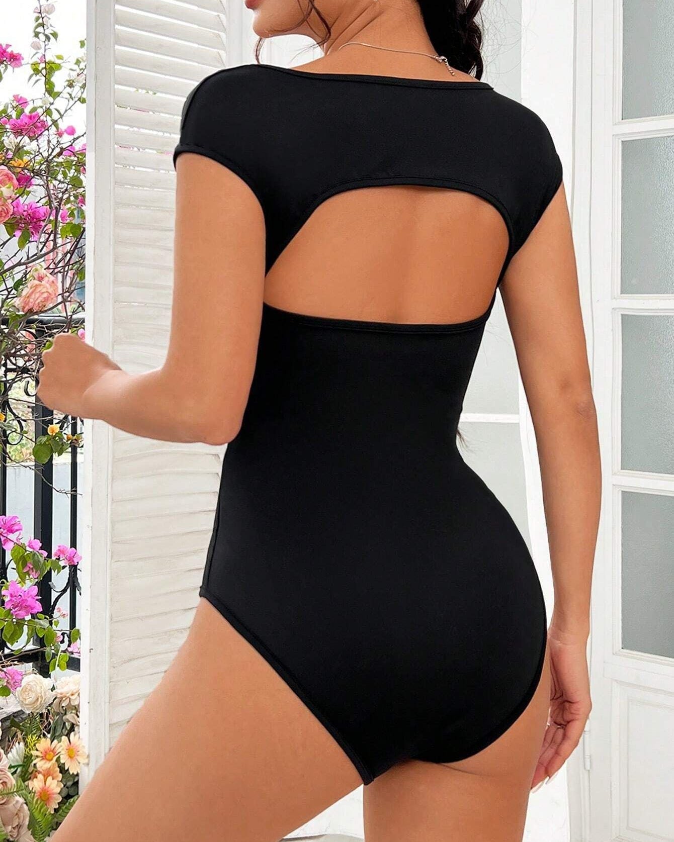Back view: Open-back black bodysuit with a curved cutout and cap sleeves, paired with floral background decor.