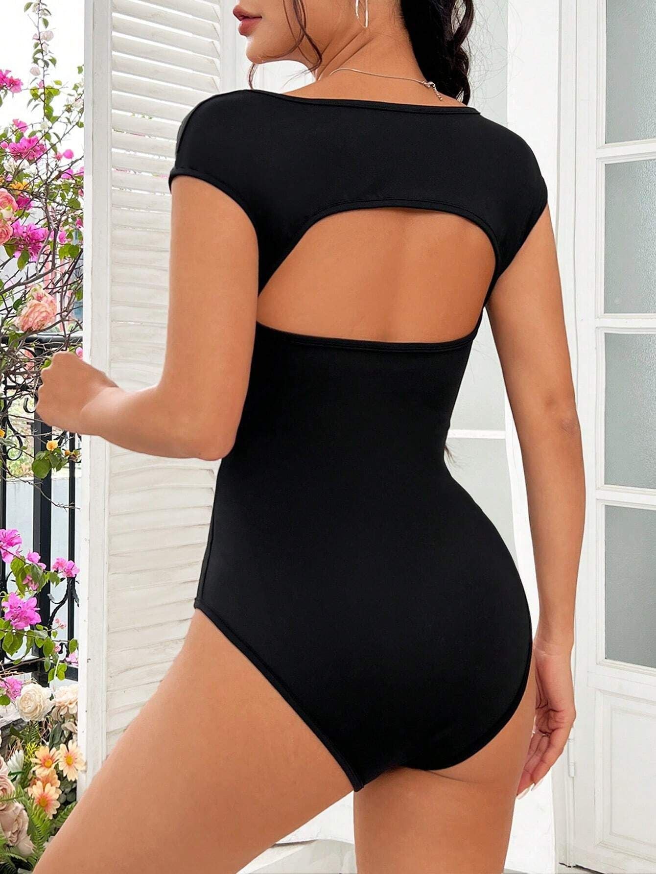 Back view: Open-back black bodysuit with a curved cutout and cap sleeves, paired with floral background decor.