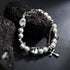 A silver chain bracelet with white pearl-like beads and metallic geometric accents, featuring a small cross pendant, displayed on a black textured surface with dark stones.