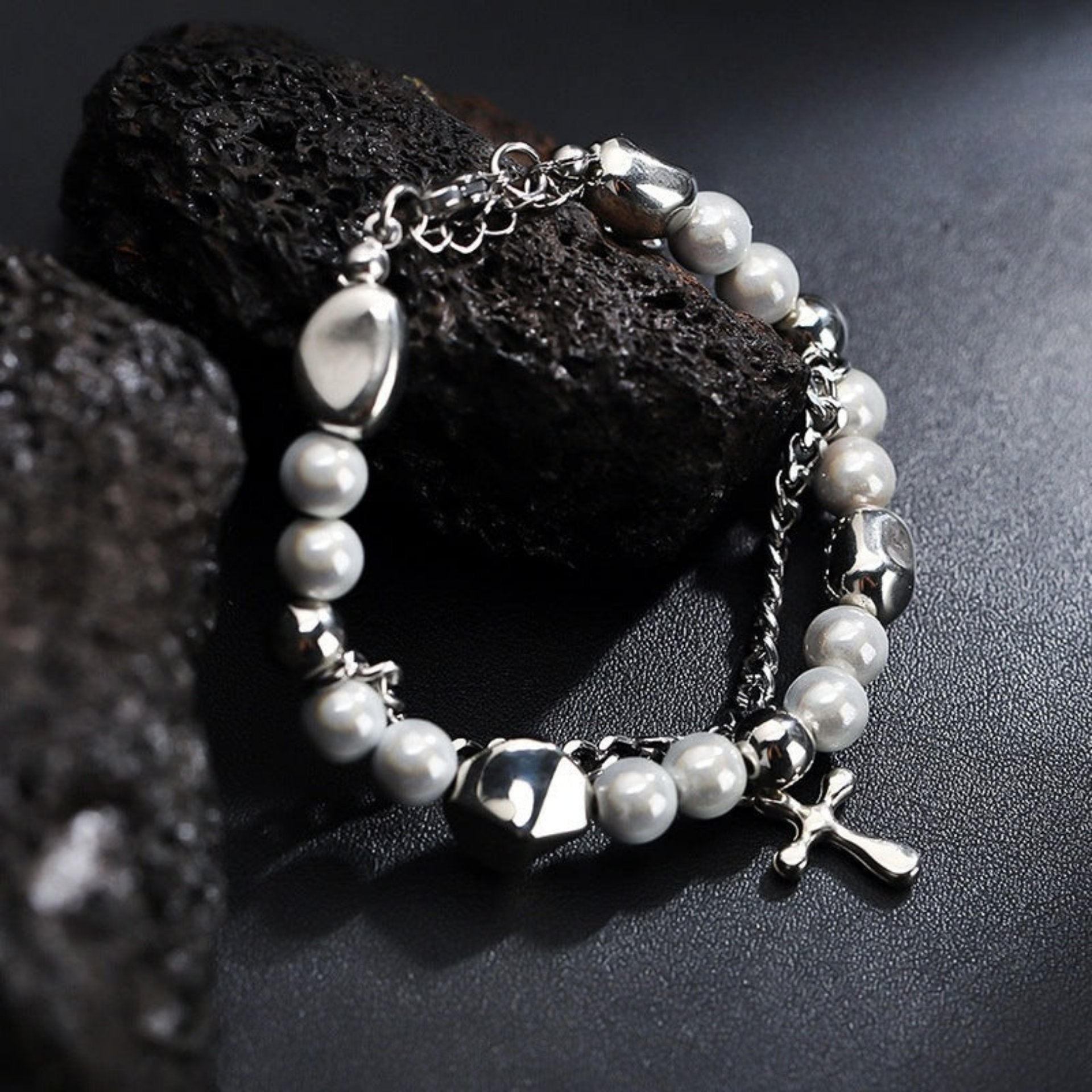 A silver chain bracelet with white pearl-like beads and metallic geometric accents, featuring a small cross pendant, displayed on a black textured surface with dark stones.