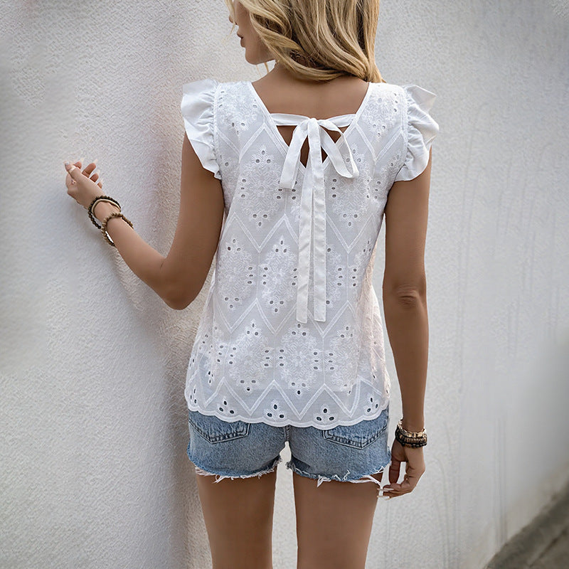 Back view of a woman in a white lace top with ruffled sleeves and a bow-tie back, paired with denim shorts, standing against a textured wall.