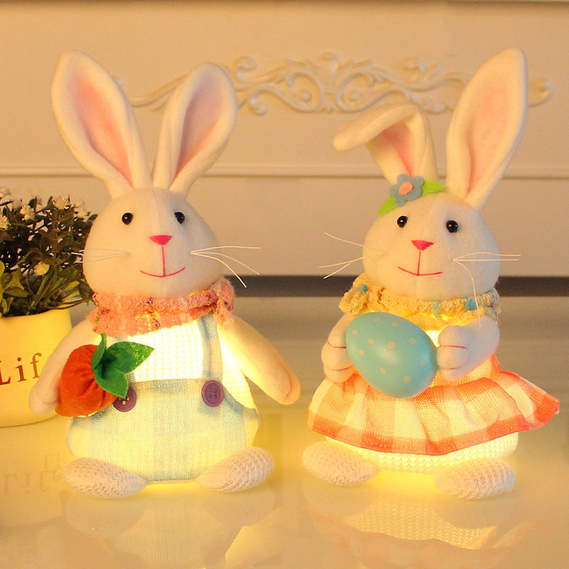  Two plush bunny decorations with glowing bellies, one in blue overalls holding a carrot, the other in a plaid dress holding an Easter egg, placed on a table.