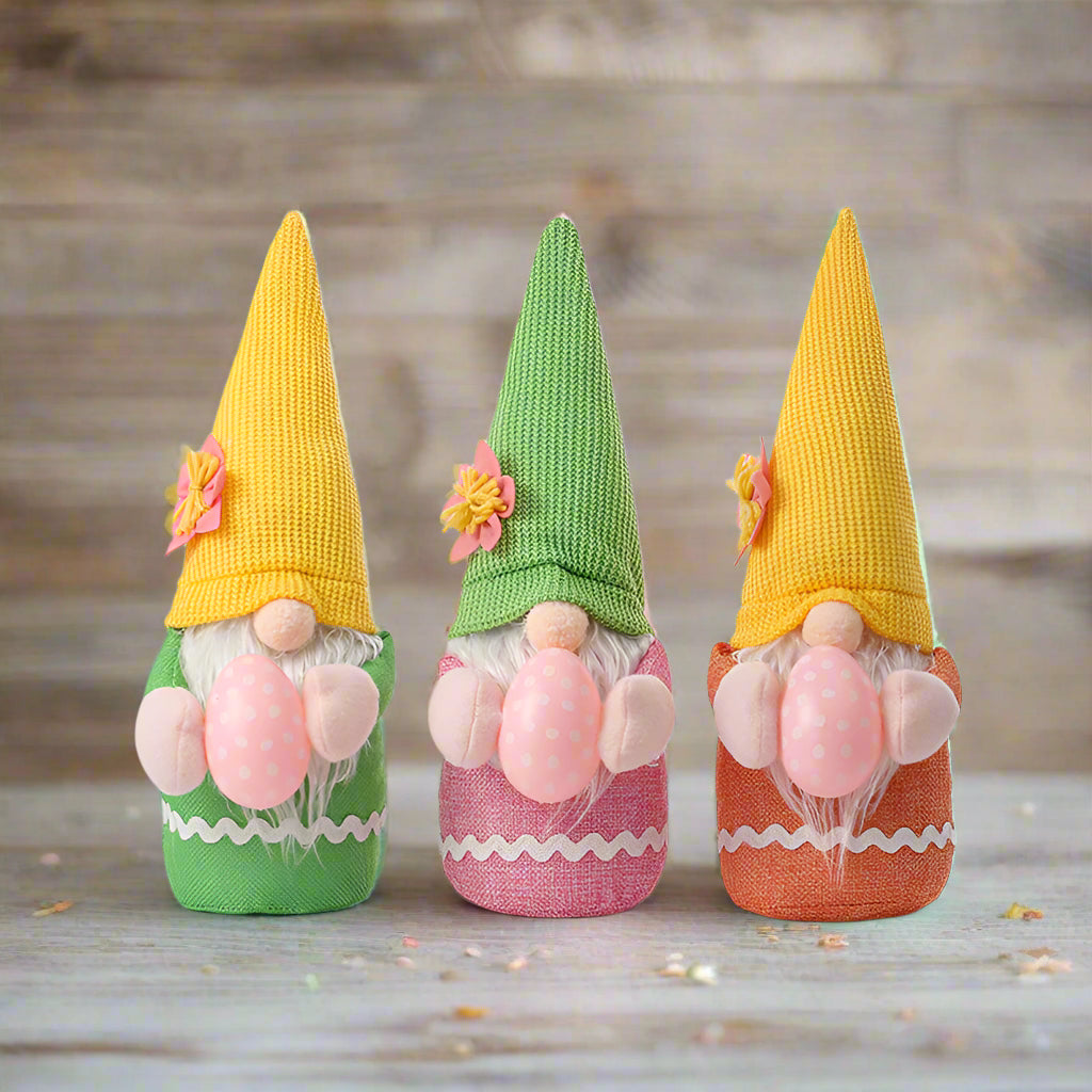 Three Easter gnome plush toys with yellow, green, and orange knitted hats, each holding a pink polka-dotted egg.