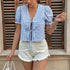 Woman wearing a blue and white gingham puff sleeve crop top with front tie details, paired with white frayed shorts, standing in front of wooden shutters.