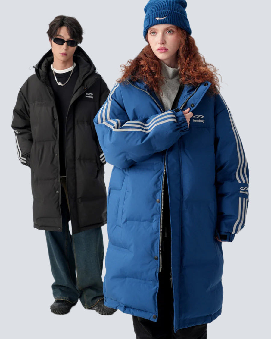 A man and woman are standing against a gray backdrop. Both wearing long puffer coats with stripe details, him in black and her in blue with matching beanie