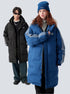 A man and woman are standing against a gray backdrop. Both wearing long puffer coats with stripe details, him in black and her in blue with matching beanie