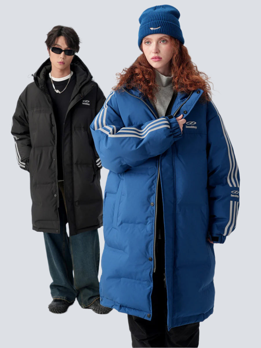A man and woman are standing against a gray backdrop. Both wearing long puffer coats with stripe details, him in black and her in blue with matching beanie