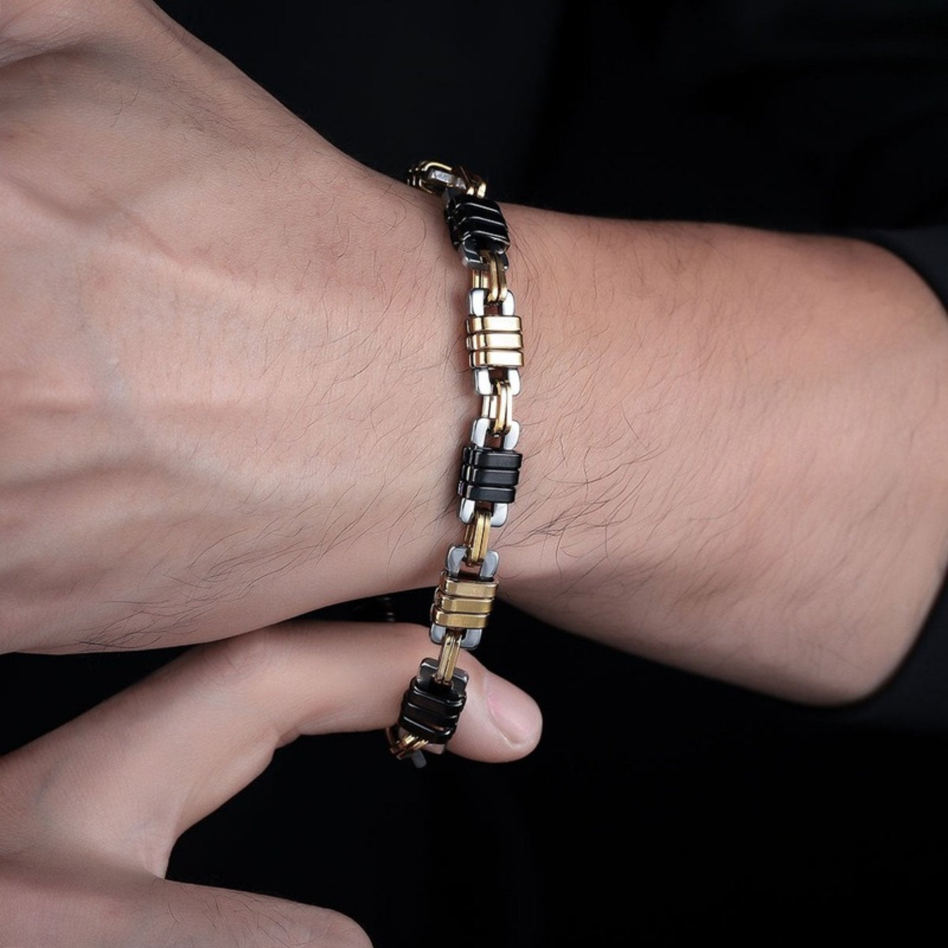  Close-up of a man's wrist wearing a black and gold stainless steel bracelet with a mechanical-inspired link design against a dark background.